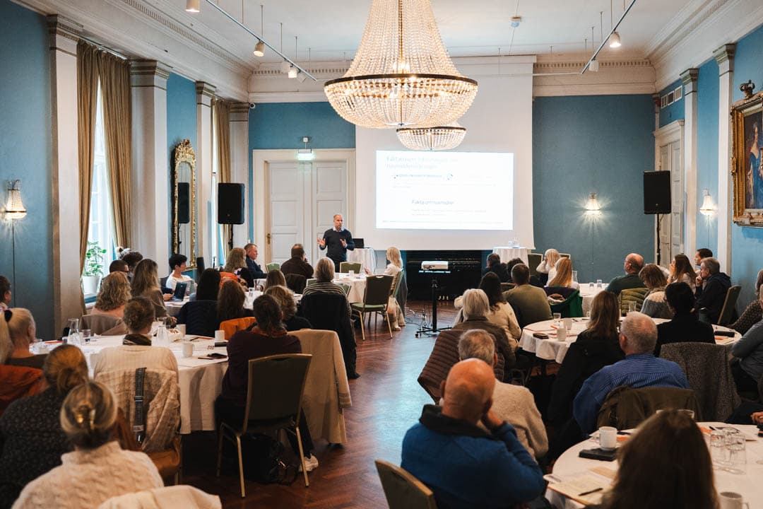 gruppe mennesker som sitter i et rom og hører på to som står framme og snakker.