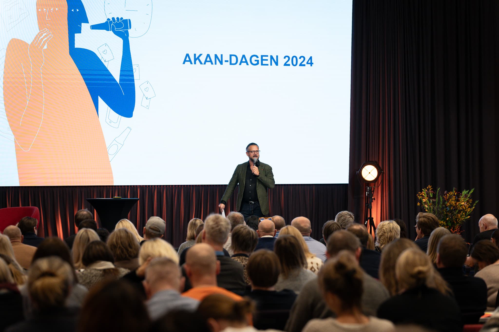 Person står på scenen foran mange folk og snakker i mikrofon. Bak ham er det en stor skjerm hvor det står "Akan-dagen 2024".