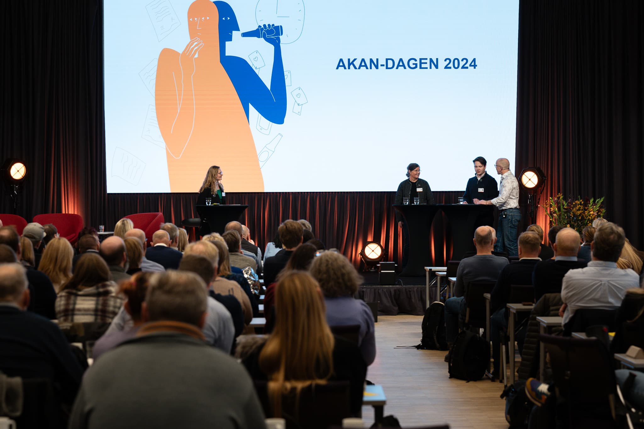 Fire personer står på scenen foran mange mennesker. På skjermen bak dem står det "Akan-dagen 2024".